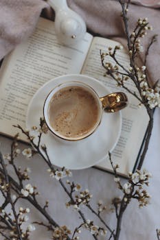 From above of mug of hot drink on plate placed on opened book on table with decorative bird and blooming branches