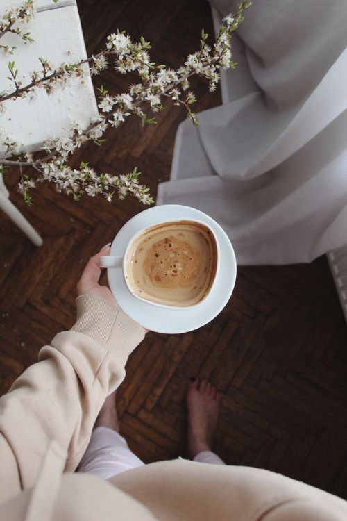 Free From above of crop faceless barefoot female with cup of coffee in hand standing near blooming branches in light room with tulle Stock Photo