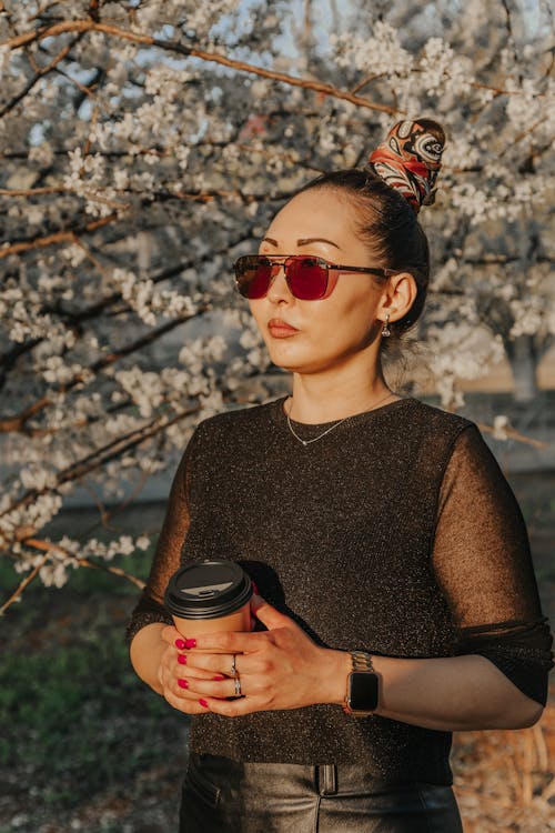 Free Cool ethnic female in sunglasses and smart watch holding takeaway hot drink while looking away under blooming tree in sunlight Stock Photo