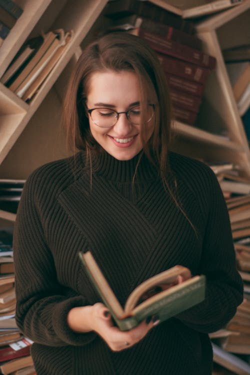 Free Cheerful female in warm black sweater reading interesting book while standing near bookshelves with various literature in modern light library Stock Photo