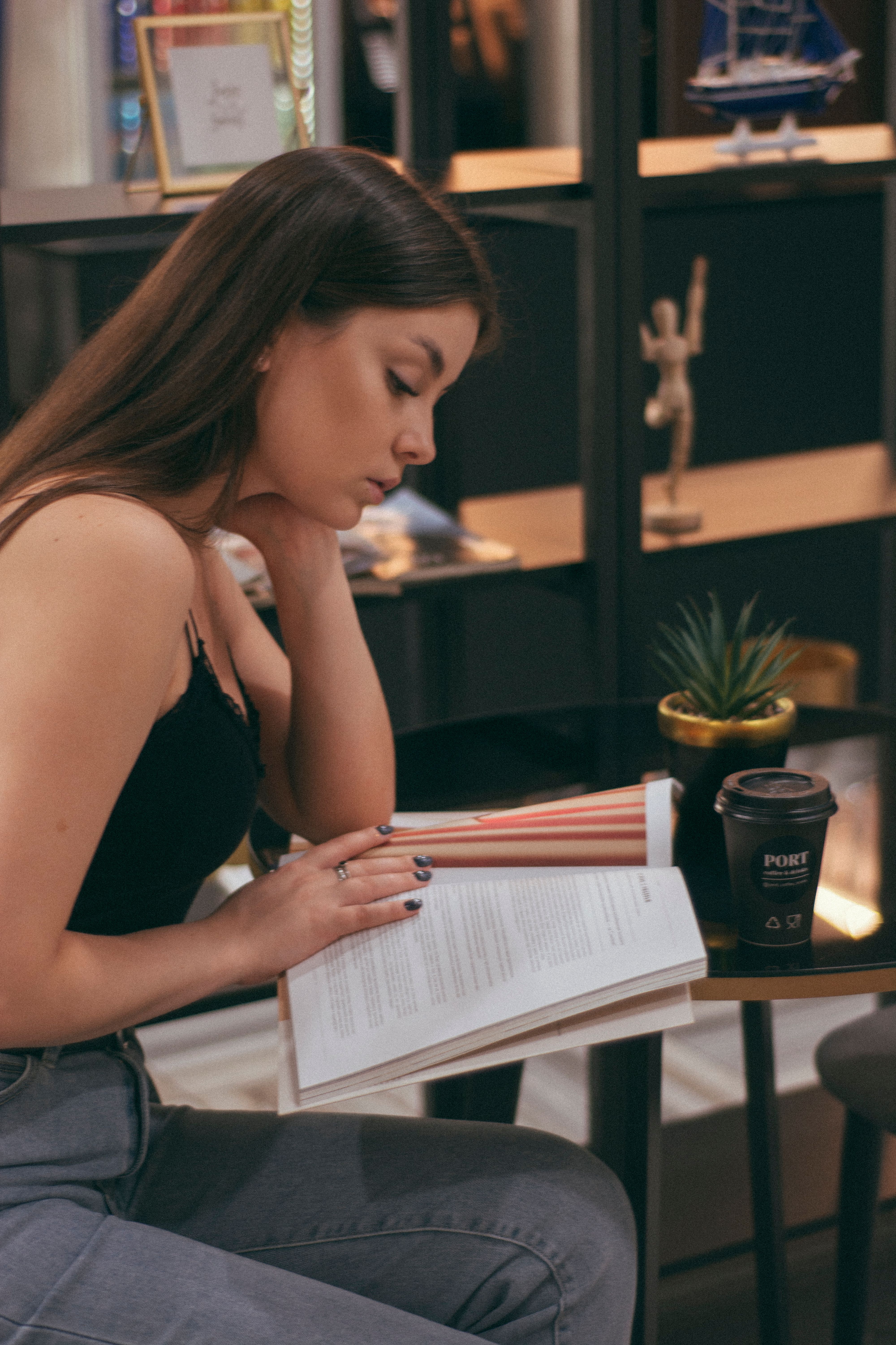 Side view of concentrated young female reading magazine while sitting at table with takeaway hot coffee near shelves with decorations