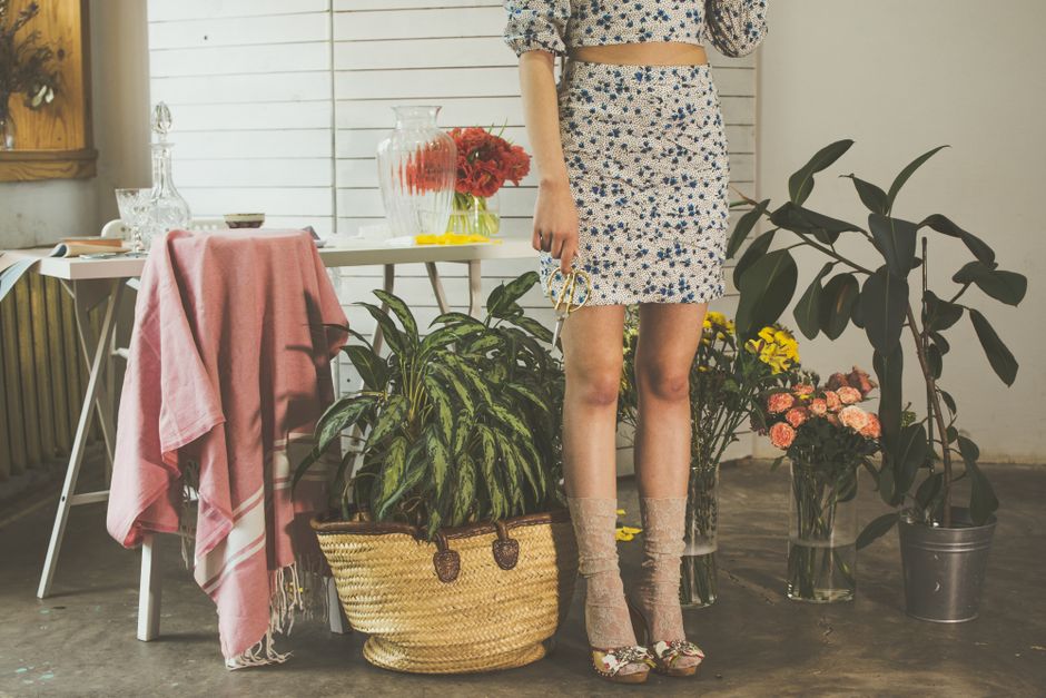 Crop anonymous female wearing skirts and high heels standing near table with vase in light room with green potted plants