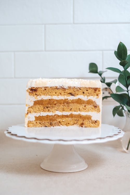 Delicious cut cake with filling and white frosting placed on stand on table with green plant in kitchen on white background