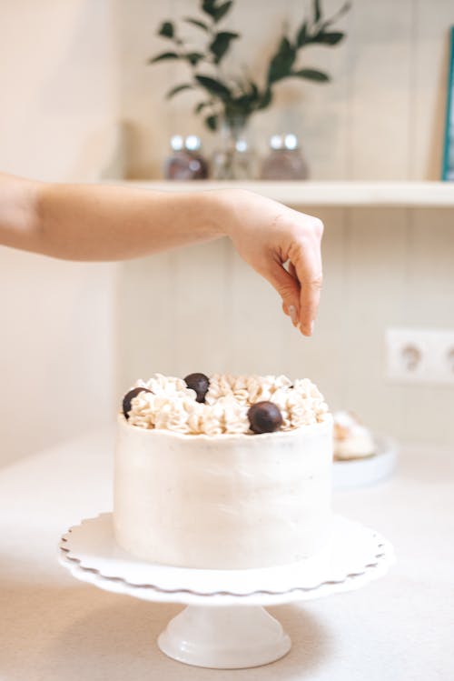 Free Crop unrecognizable person sprinkling sweet decorated cake with white frosting and candies served on stand on table in light kitchen Stock Photo