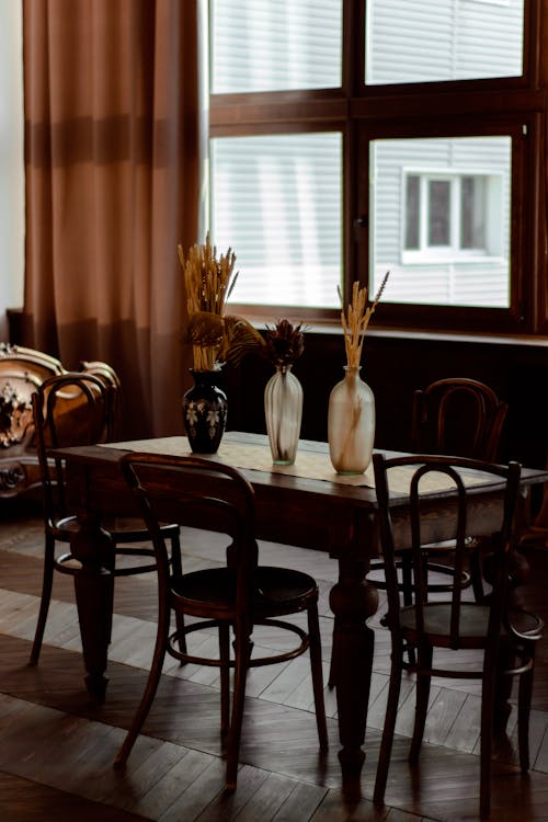 Different vases with various dried flowers placed on wooden table with chairs in modern light room with curtain on window