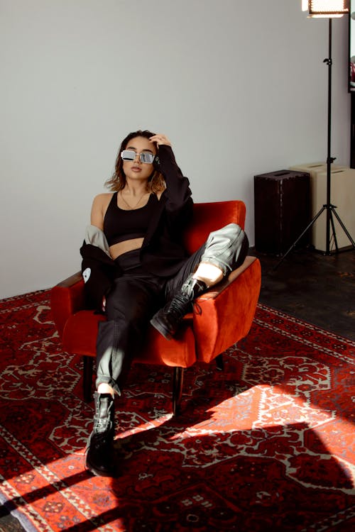 Full body of young confident female in trendy clothes and sunglasses sitting in armchair on carpet in light apartment