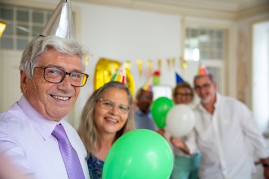 Seniors joyfully celebrating a birthday indoors with balloons and party hats.