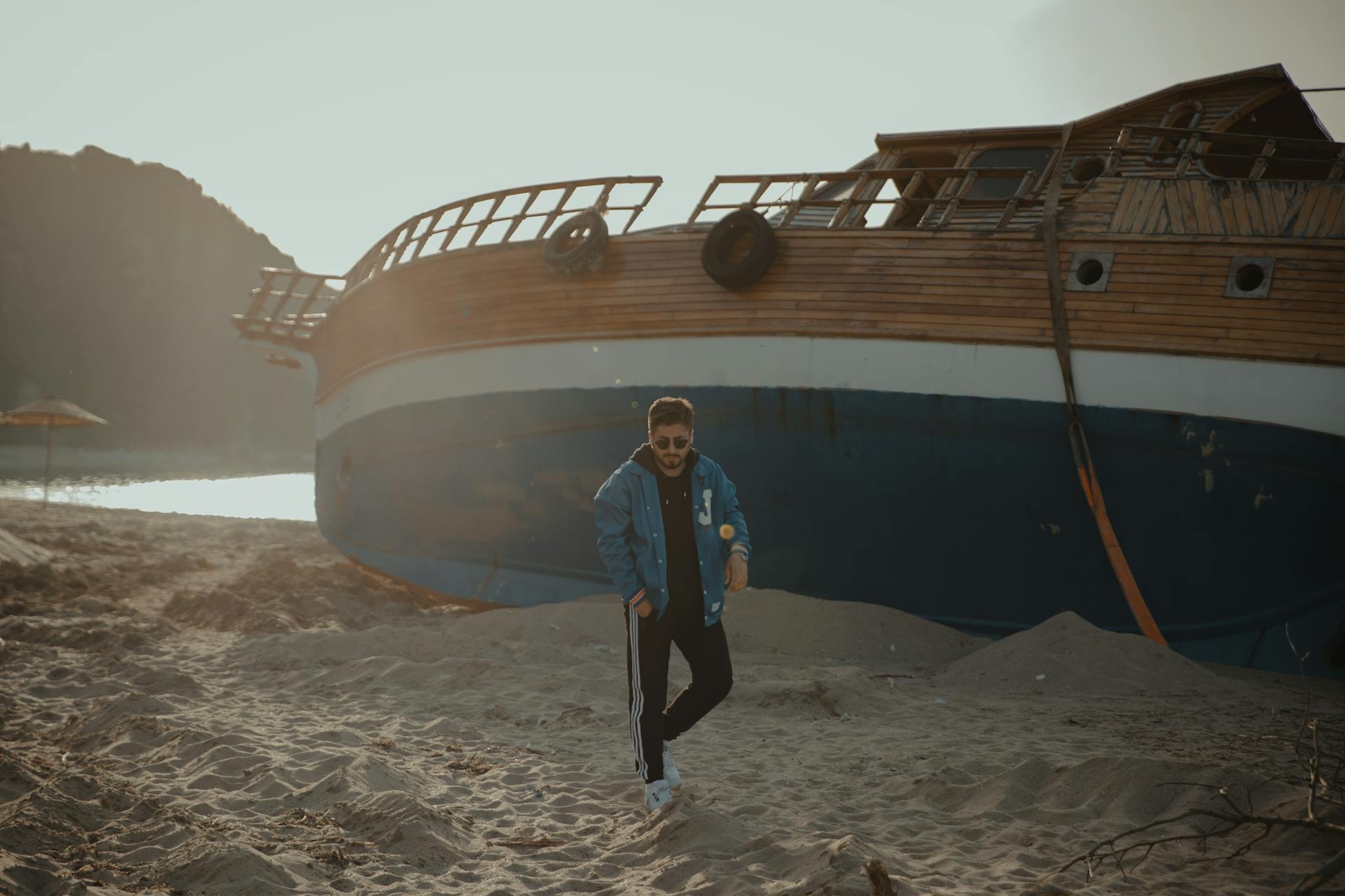 A man walks along a sandy beach with a shipwreck in the background during sunset.