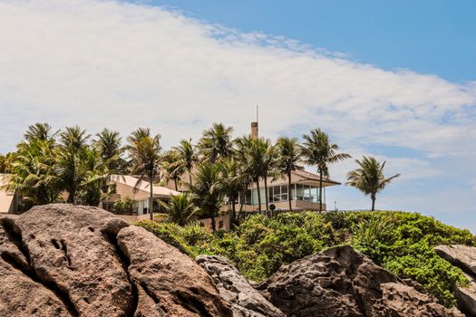 A stunning tropical villa surrounded by palm trees on a rocky coastline under a bright sky.