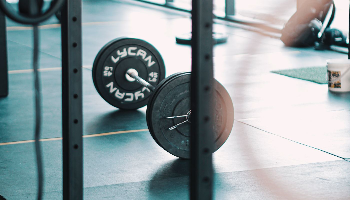 A loaded barbell resting on the floor of a daylit gym, seen between the black uprights of a rack, with a figure out of focus in the background