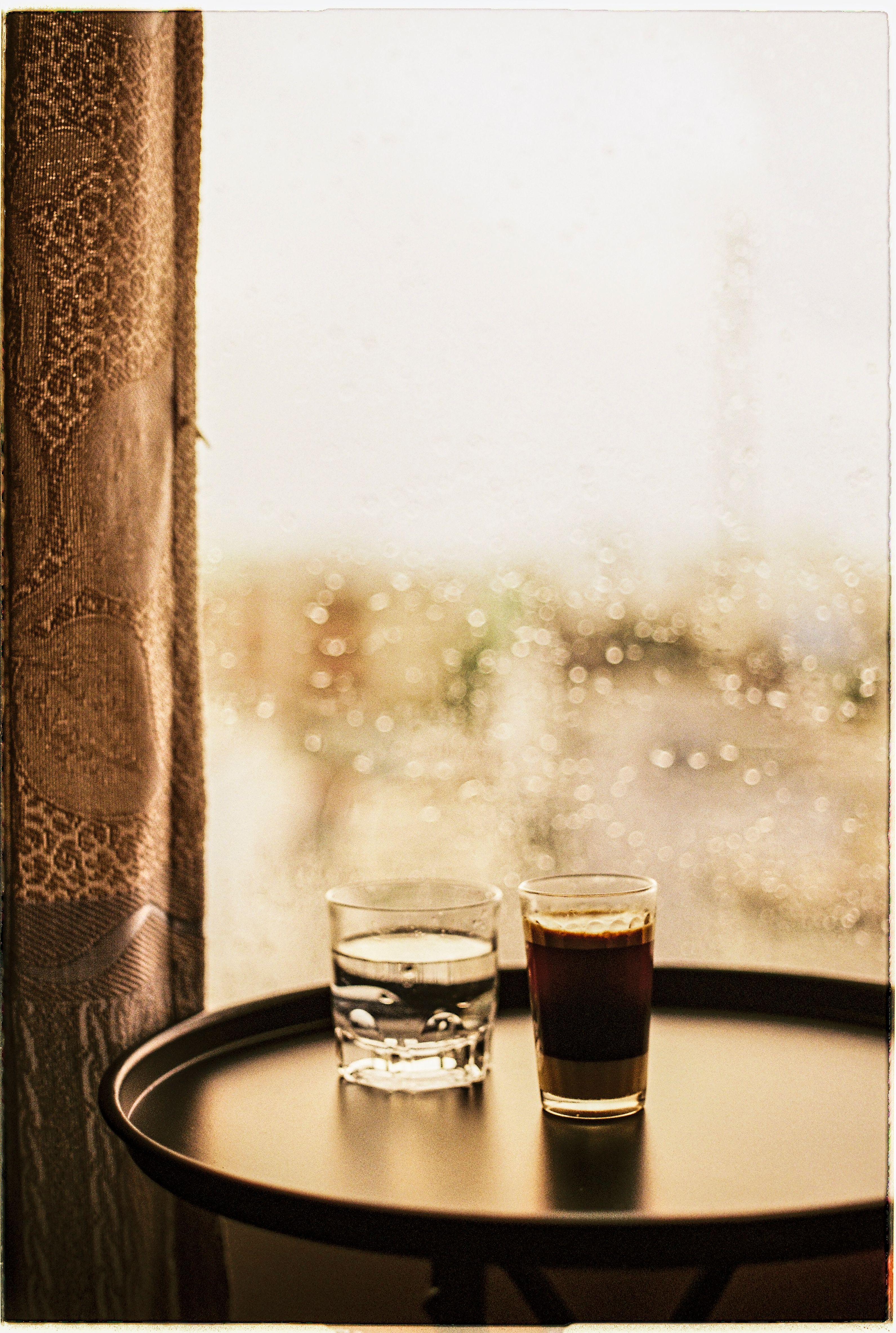 Cozy morning by the window with coffee and water, capturing raindrops and warmth.