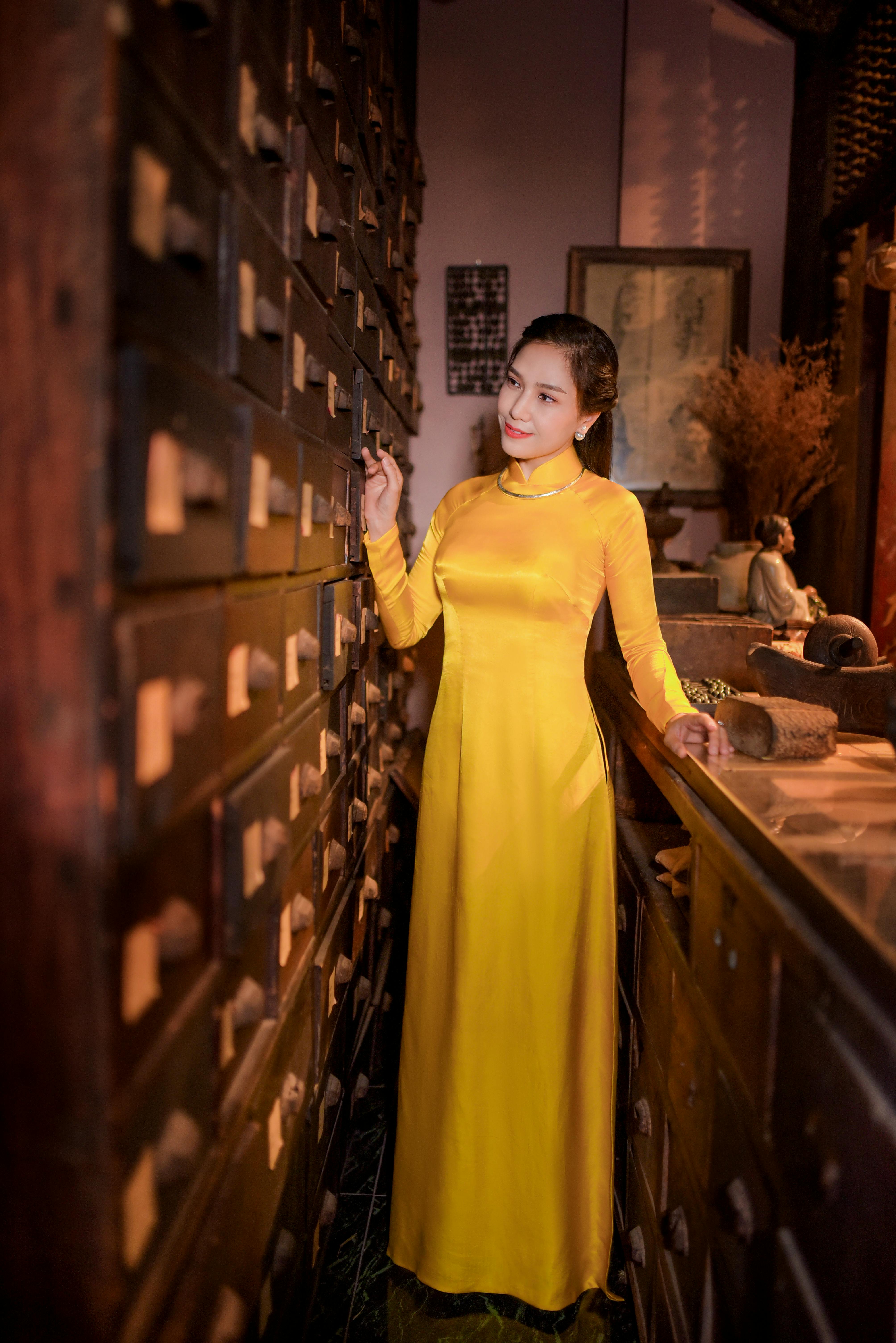 A woman in a yellow ao dai stands gracefully in an antique room filled with drawers.
