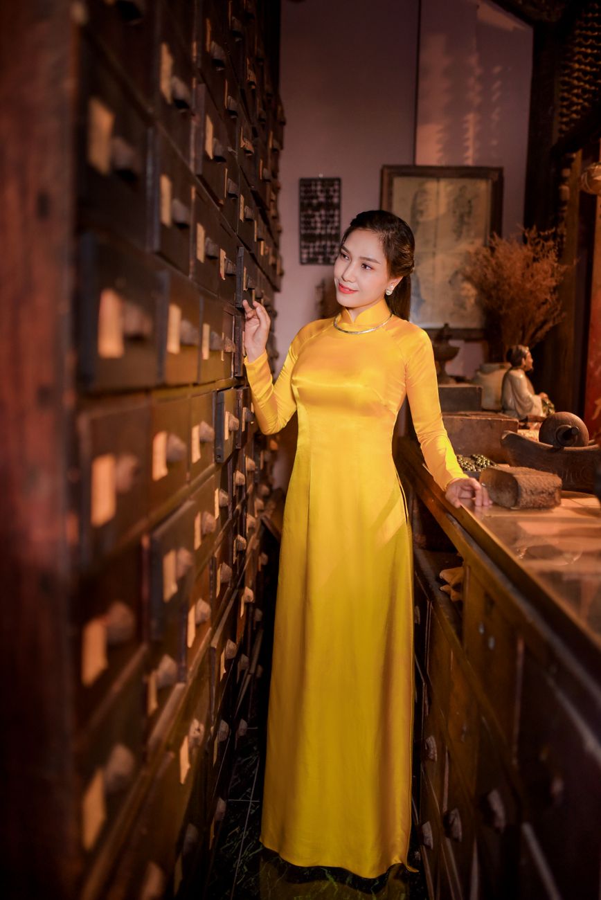A woman in a yellow ao dai stands gracefully in an antique room filled with drawers.
