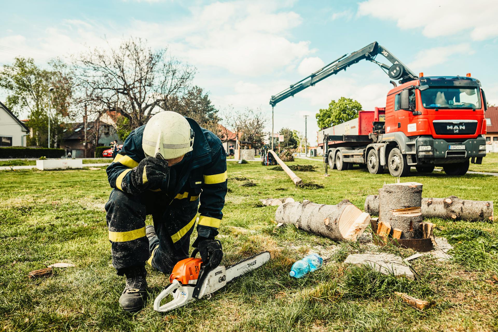 Arborist in safety gear using chainsaw beside truck with crane in an outdoor setting.