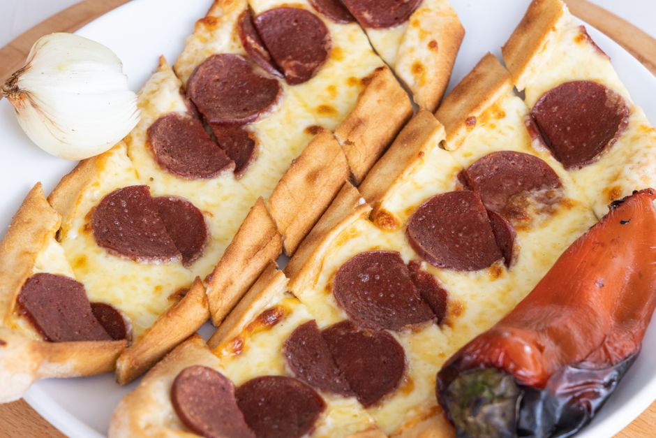 Flattened dark red-brown sujuk sausage sliced on a diagonal into thin ovals, some pieces frying in a skillet alongside cracked eggs.