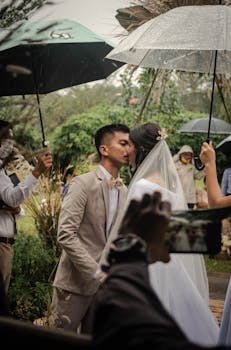 Bride and groom share a kiss under umbrellas during a rainy outdoor wedding.