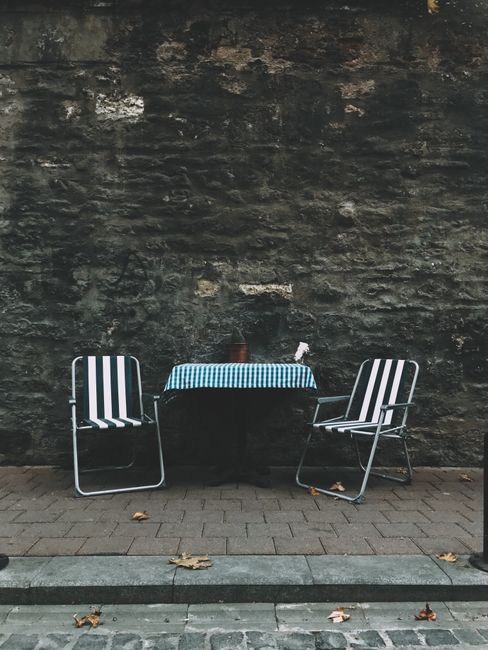 Beach chairs near table with tablecloth placed on paved sidewalk with fallen leaves at shabby stone wall on street of town