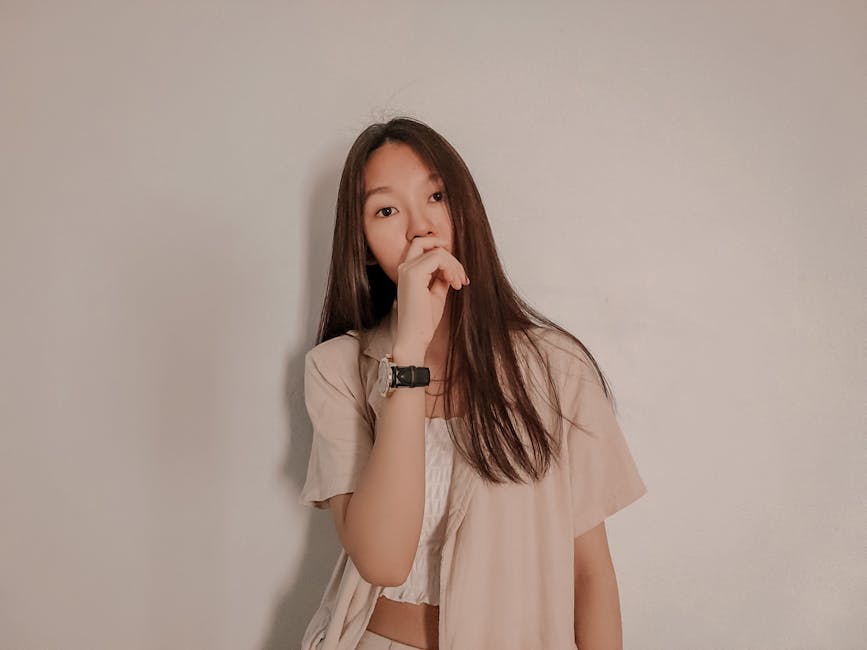 Young woman in casual outfit stands against a plain white wall, looking thoughtful.