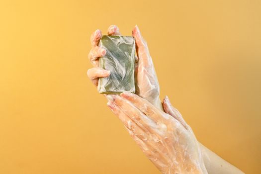 Hands covered in foam holding a green bar soap against a yellow background, promoting hygiene.