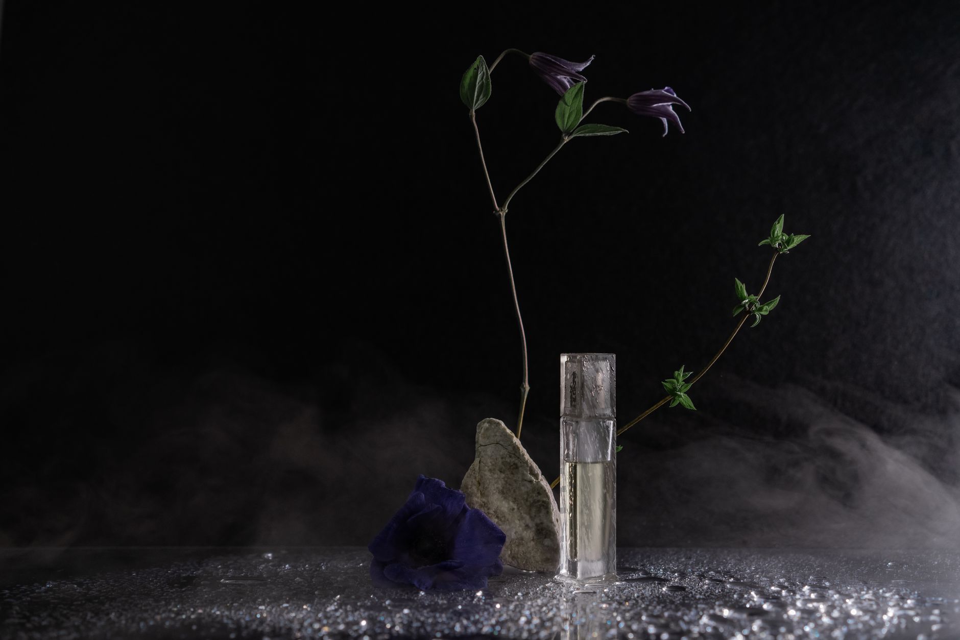 Artistic still life featuring purple flowers, rock, and glass perfume bottle against black background.