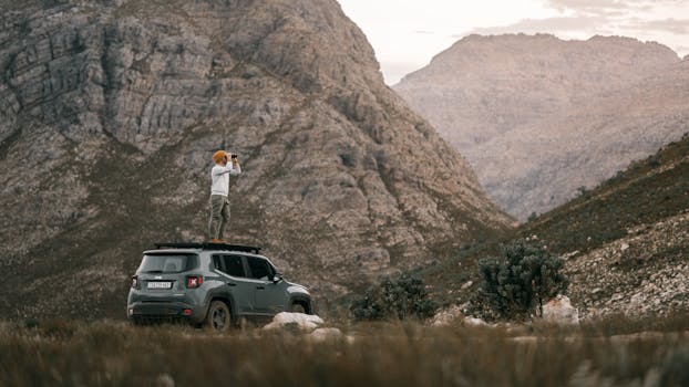 A man standing on an SUV roof using binoculars to view a mountain landscape.