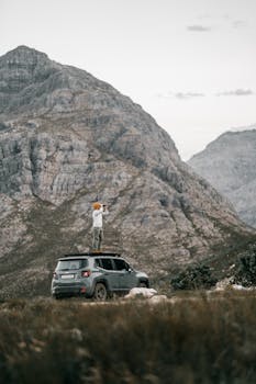 A person stands on a SUV using binoculars, exploring rocky mountains in an overcast setting.