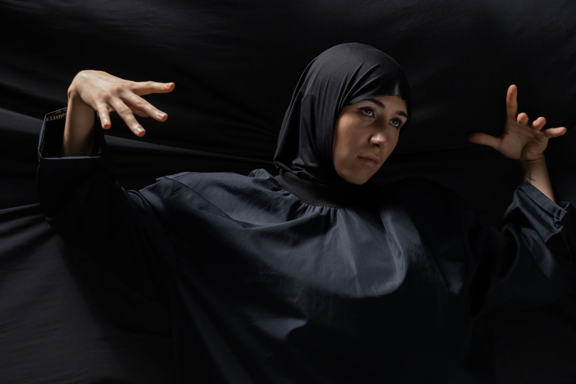 Portrait of a woman in black traditional clothing and hijab posing with expressive hands against a dark background.