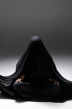 Mysterious person draped in black fabric seated in a studio setting, creating an enigmatic silhouette.
