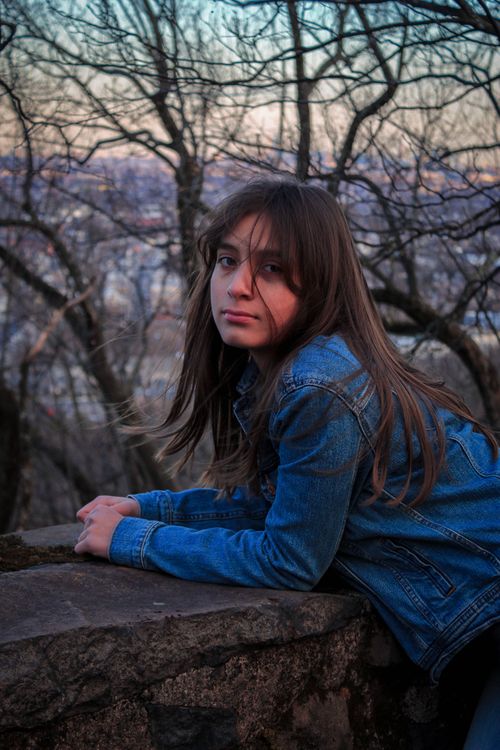 Free A young woman in a denim jacket leans on a stone wall amidst leafless trees in autumn. Stock Photo