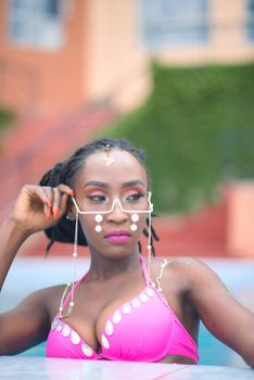 Fashionable black woman posing in a pool wearing stylish eyewear and a bright bikini.