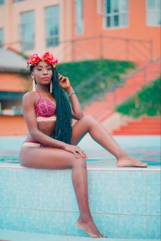 Beautiful African woman poses poolside in a bikini, exuding confidence with long hair and floral headpiece.