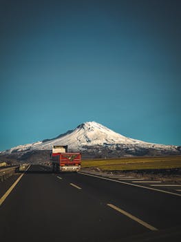 A truck travels on a rural road towards a scenic snow-covered mountain under a clear blue sky.