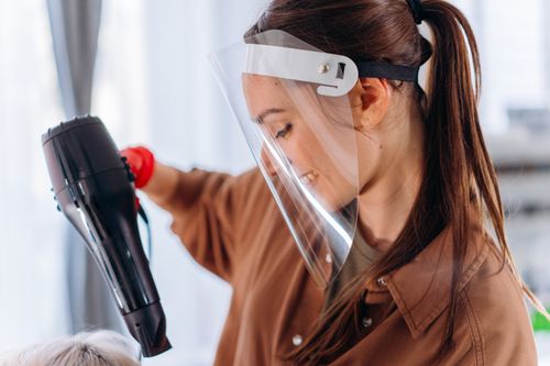 Free Hairdresser wearing a face shield, using a hair dryer to style hair. Indoor salon setting with a safety focus. Stock Photo