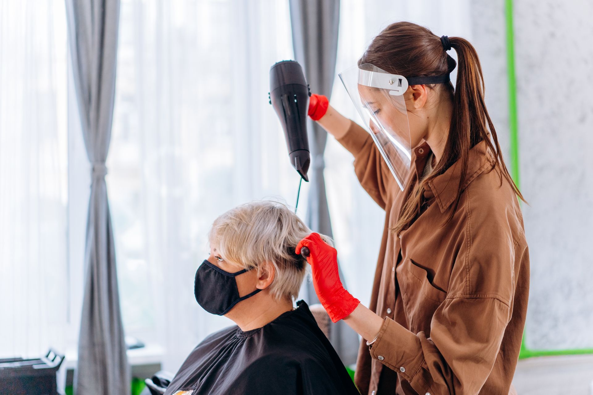 A hair stylist wearing a face shield and gloves dries a client's hair in a salon setting.