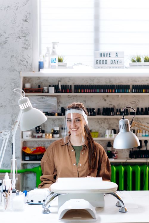 Free A cheerful manicurist wearing a face shield smiles in a well-equipped modern nail salon. Stock Photo