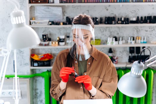Manicurist wearing face shield and gloves, preparing nail polish in a clean, indoor salon environment.