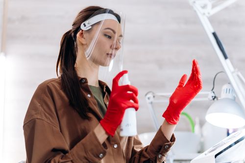 Free Manicurist wearing a face shield and red gloves in a salon, holding a spray bottle as part of safety measures. Stock Photo