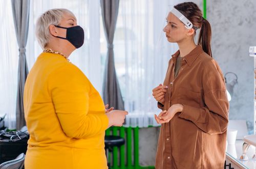 Free Two women conversing indoors, one wearing a face mask and the other a face shield, reflecting pandemic safety measures. Stock Photo
