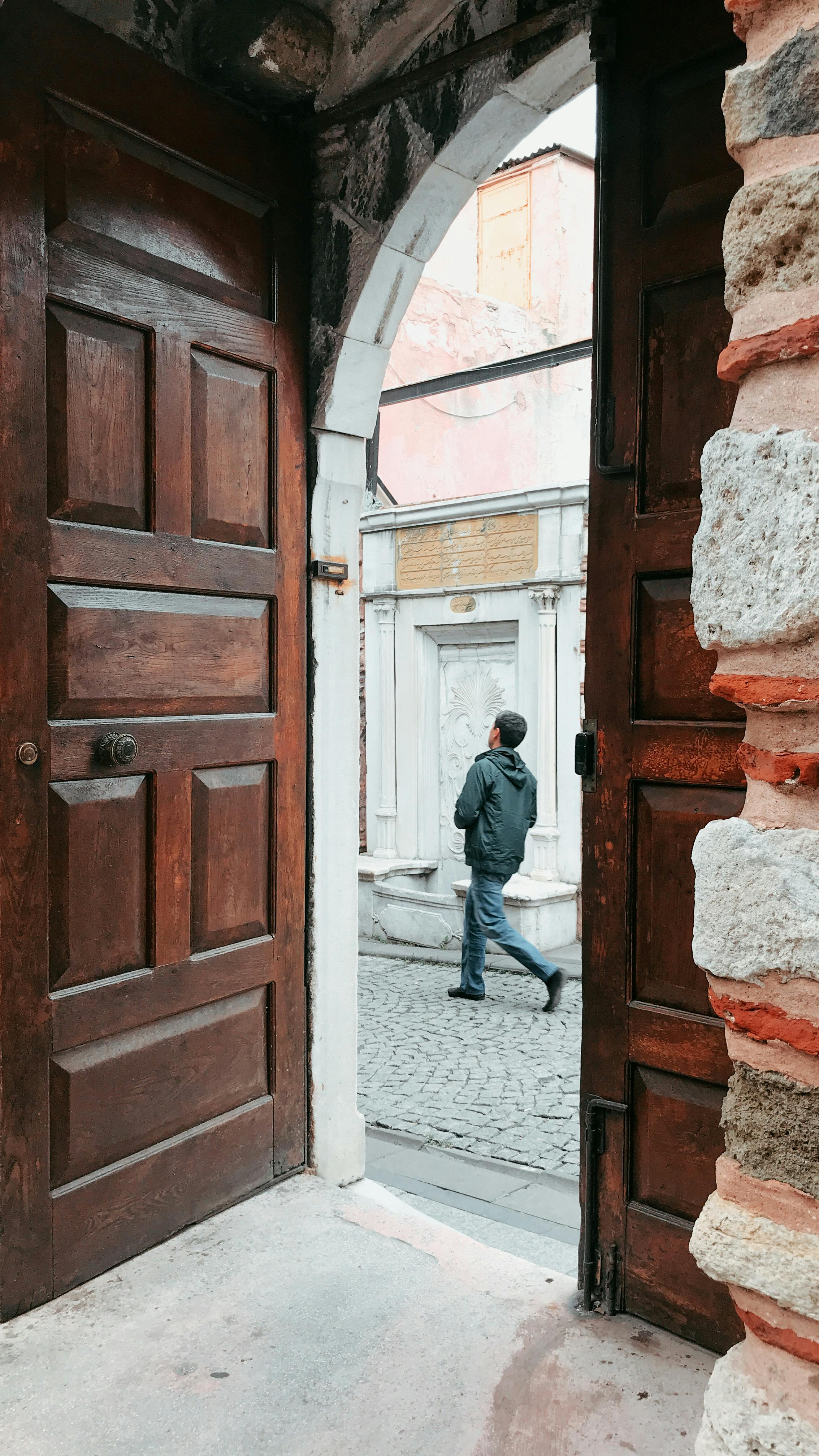 Opened wooden door of aged stone house overlooking distant faceless male strolling on paved sidewalk on street in old town