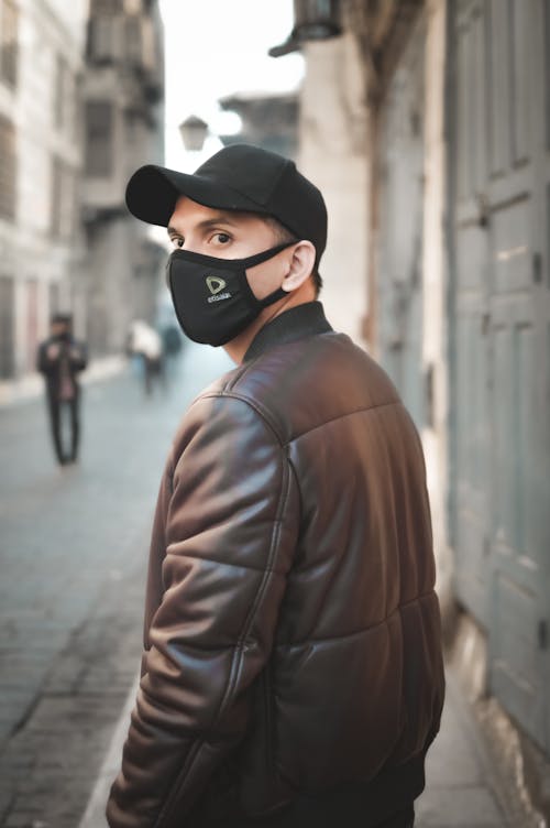 A Man in Face Mask and Leather Jacket Walking at the Sidewalk