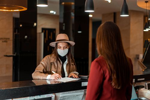 Free A woman in a hat and face mask checks in at a hotel reception, interacting with a receptionist. Stock Photo