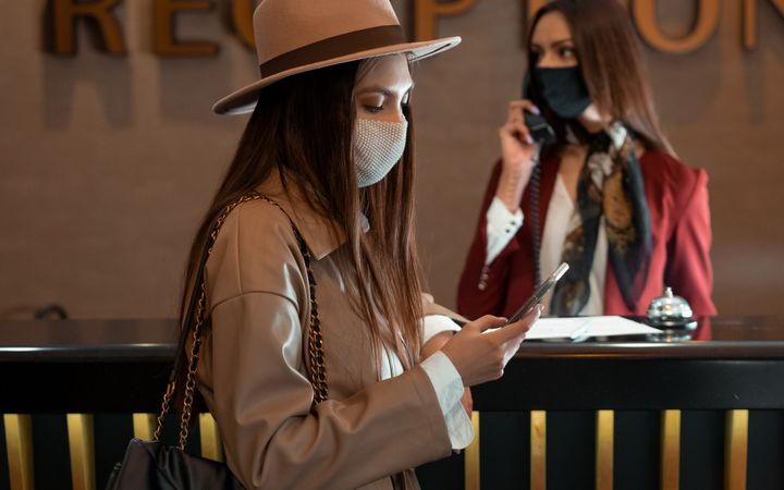 Woman in face mask at hotel reception counter checking her phone, symbolizing the new normal.