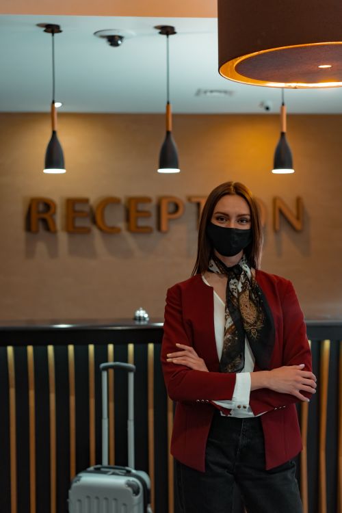 Free A stylish woman wearing a mask stands at the hotel reception, arms crossed, near luggage. Stock Photo