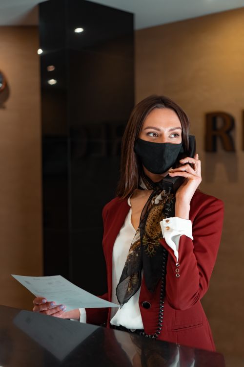 Free A professional receptionist in a red blazer talks on the phone at a hotel front desk, holding documents. Stock Photo