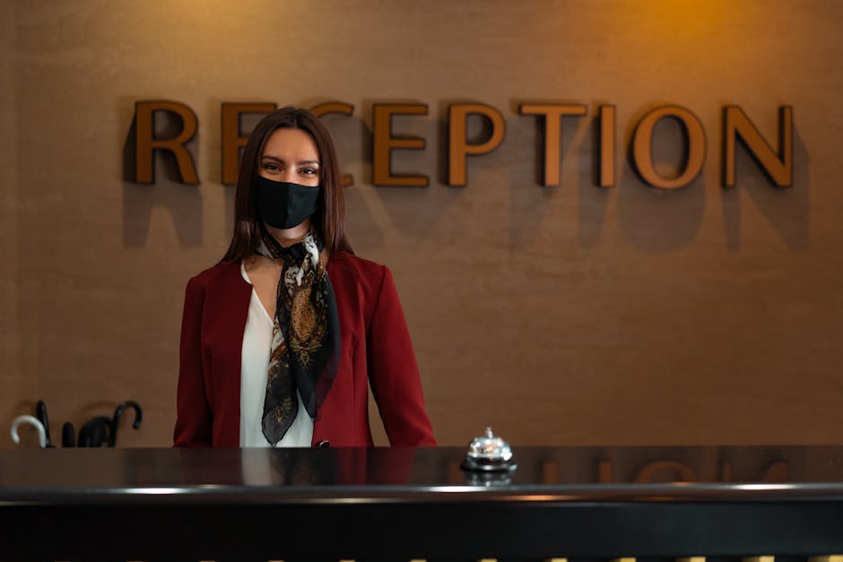 Woman in mask at hotel reception desk ready to assist guests.