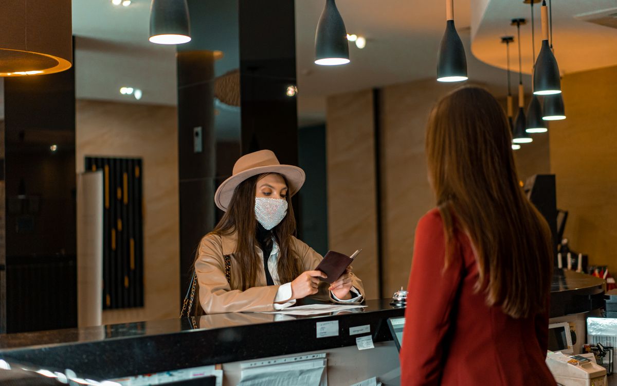 A woman wearing a mask checks in at a hotel reception desk.