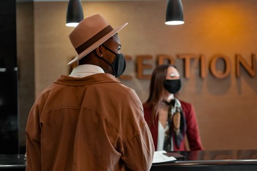 Free Man in mask at hotel reception during pandemic, interacting with masked receptionist. Stock Photo