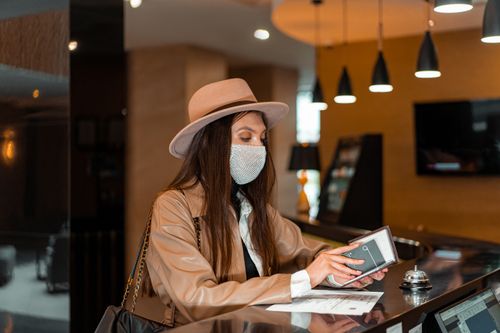 Free Woman wearing mask and hat checking in at hotel front desk, symbolizing travel and safety. Stock Photo
