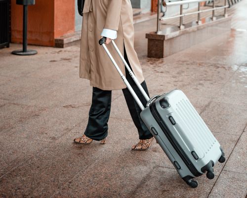 Free A person in a brown coat walking with rolling luggage on a wet pavement. Stock Photo