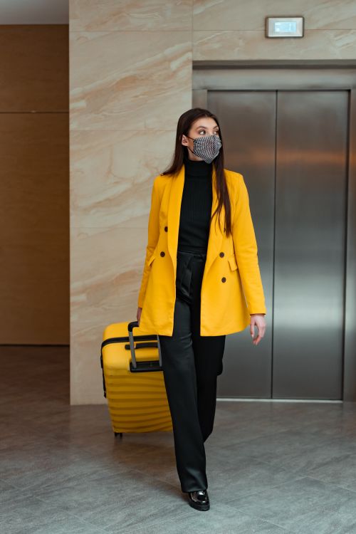 Free Woman in yellow coat with suitcase and mask walking in modern hotel lobby. Stock Photo
